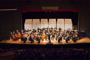 A large orchestra performs on a concert hall stage, with a conductor standing at center facing seated musicians and an audience visible in the foreground.
