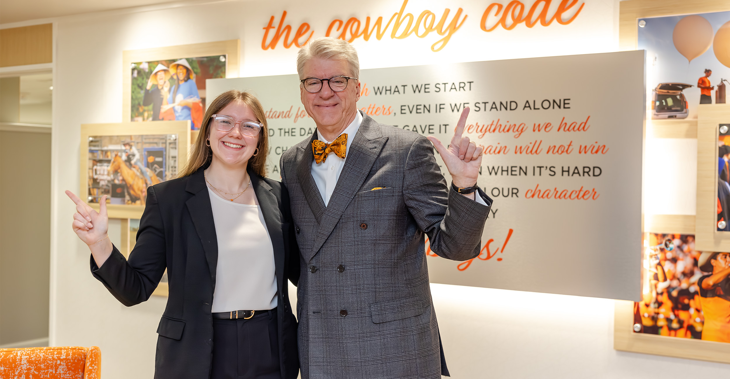 Two people stand indoors in front of a wall display titled “the cowboy code,” posing together with hand gestures in a professional setting that highlights campus values and community messaging.