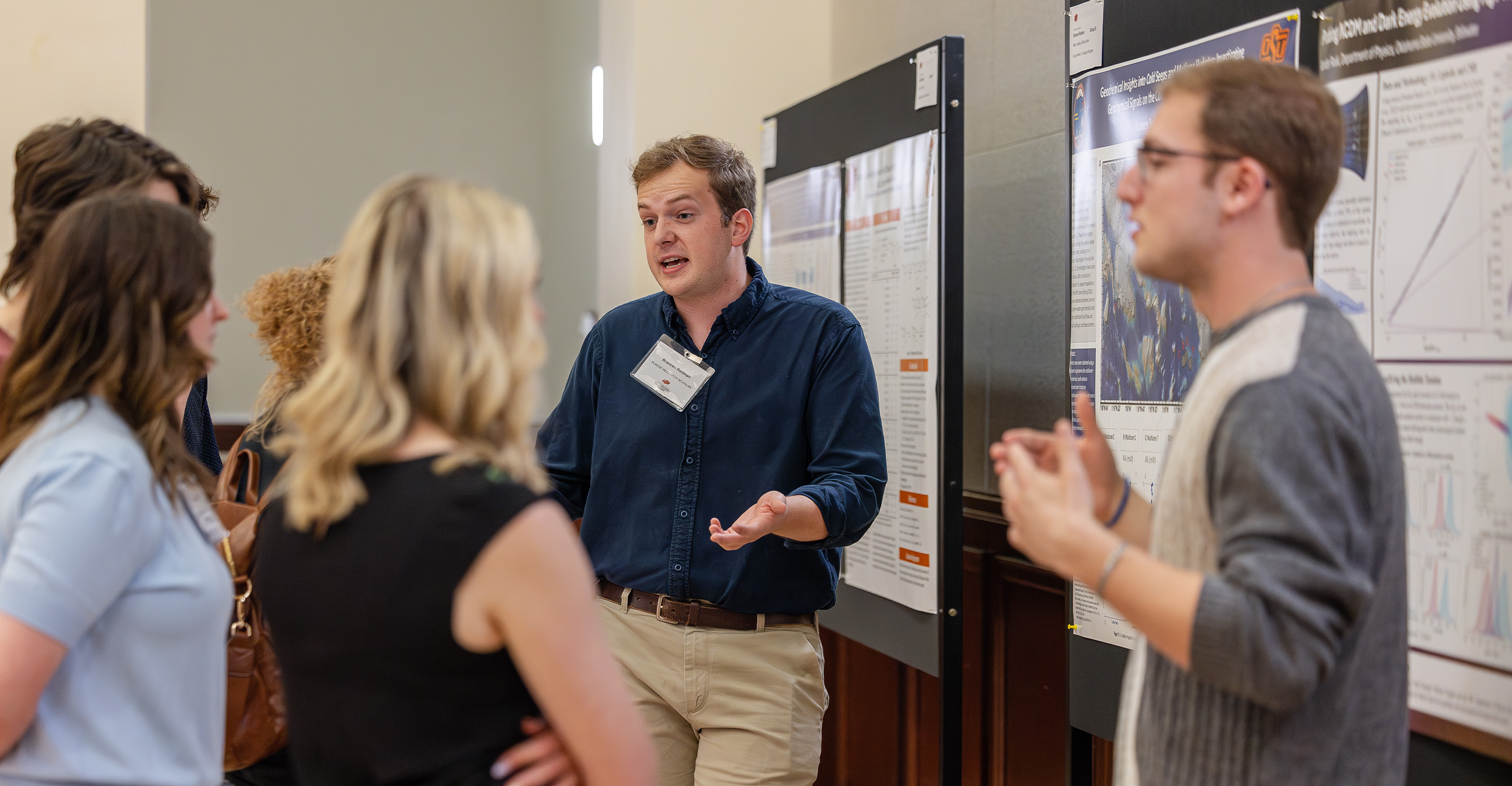 A person stands beside poster boards and gestures while explaining research findings to a small group gathered at a conference-style poster presentation session.