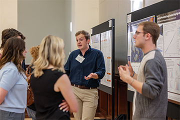 A person stands beside poster boards and gestures while explaining research findings to a small group gathered at a conference-style poster presentation session.