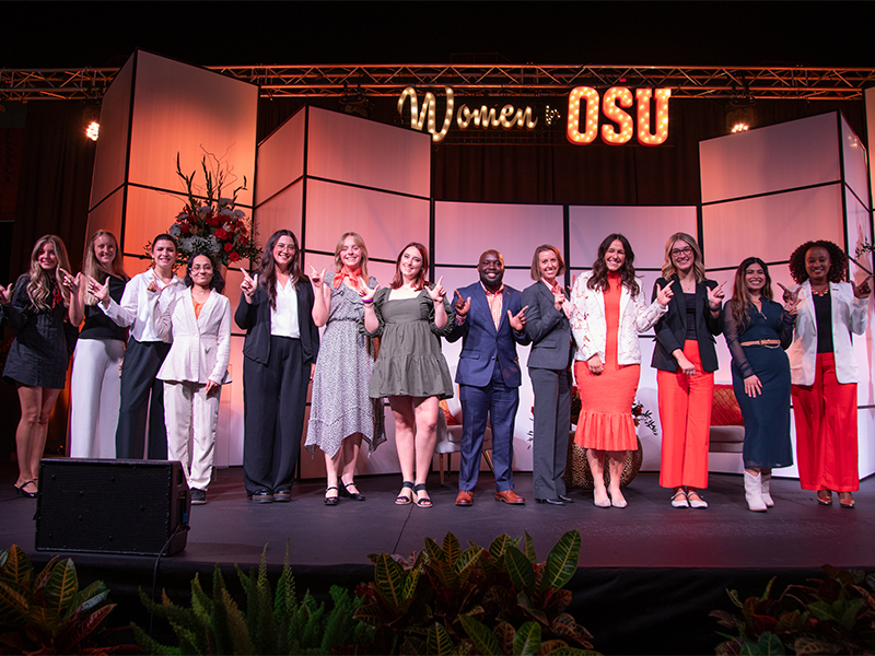 A group of people stand on a stage beneath illuminated signage reading “Women for OSU,” posed for a formal group photo at a university event with decorative backdrop panels and stage lighting.