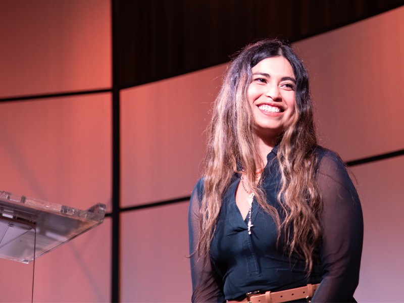 A person stands on a stage near a clear podium against a softly lit geometric backdrop, captured during a speaking or presentation moment at a Women for OSU event.