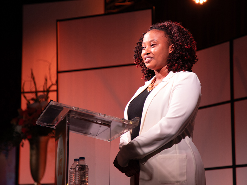 A person stands behind a clear podium on a stage with warm lighting and a geometric backdrop, delivering remarks during a Women for OSU event.