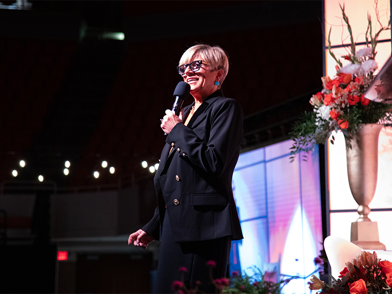 A person stands on a stage holding a microphone beside floral arrangements and stage lighting, speaking to an audience during a Women for OSU event.