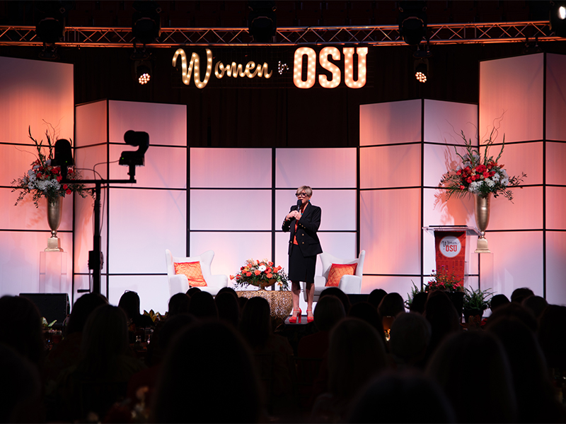 A wide view shows a speaker standing on a stage beneath illuminated “Women for OSU” signage, with decorative panels, seating, and floral arrangements visible as an audience listens in the foreground at a university event.