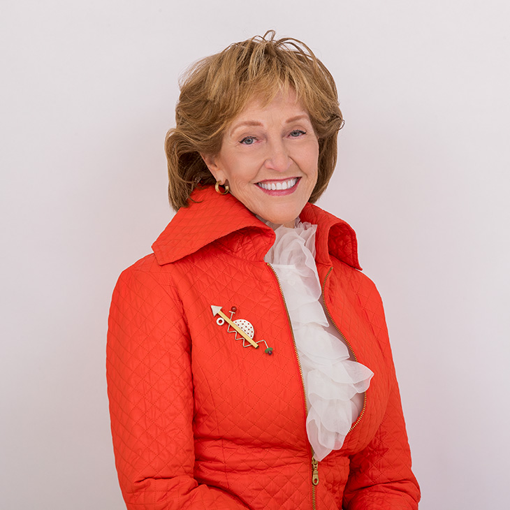 Portrait of a person wearing an orange jacket and white blouse, smiling against a plain background, highlighting a formal individual headshot.