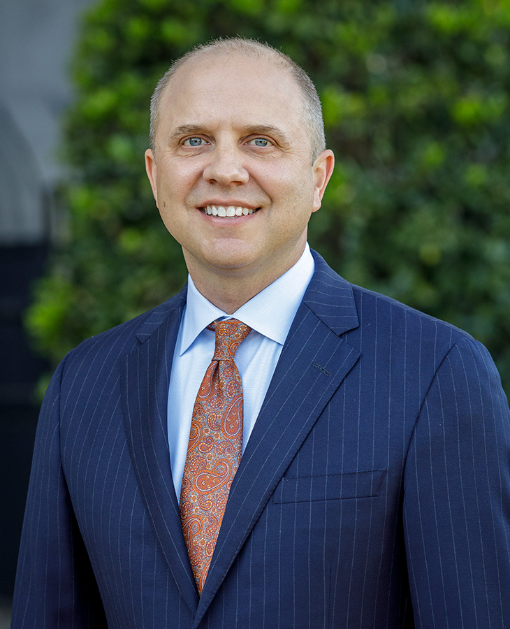 A man in a dark blue pinstripe suit, light blue dress shirt, and orange paisley tie stands in front of a background of blurred greenery.