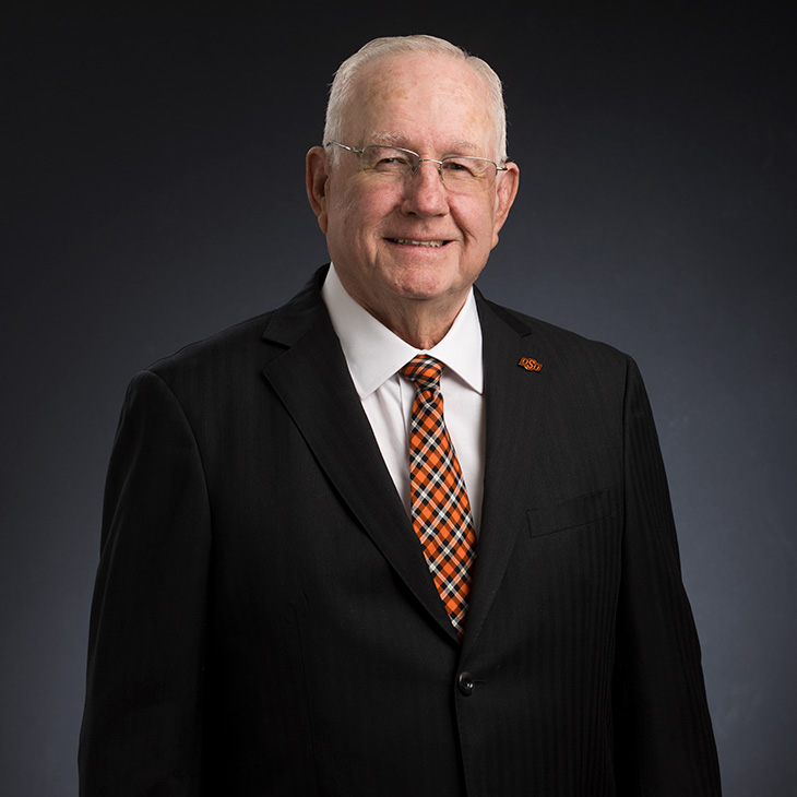 Formal studio portrait of an individual wearing glasses, a dark suit, and an orange-and-black patterned tie, posed against a dark blue gradient background.