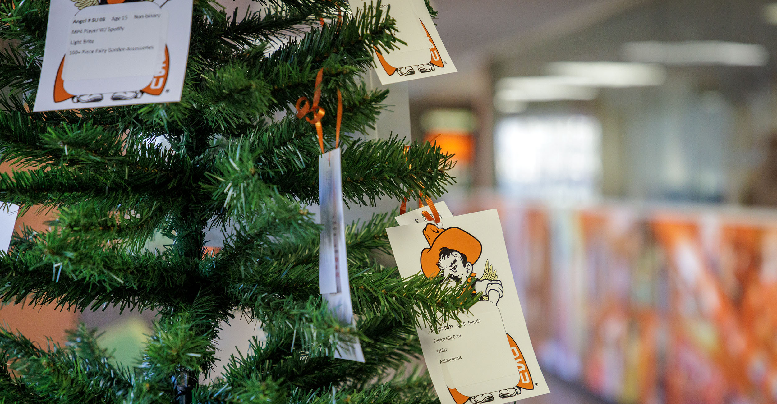 A green artificial Christmas tree indoors with several white cards hanging from its branches. The cards feature printed text and an illustration of Pistol Pete, the Oklahoma State University mascot. The background shows a blurred office hallway with orange and white decor.