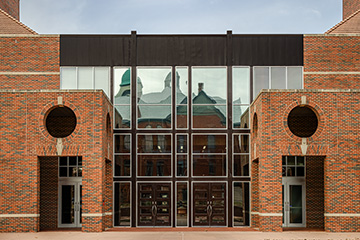 Brick building façade with large circular brick insets on each side and tall central glass windows reflecting the rooftops of nearby campus buildings.