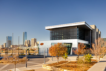 Modern glass and steel building with OSU logo in the foreground, landscaped walkway and trees, and a city skyline with tall buildings in the background under a clear blue sky.