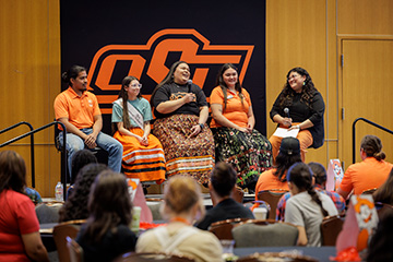 Panel of five speakers seated on a stage in front of an OSU logo backdrop, speaking into microphones during a campus event as an audience watches.