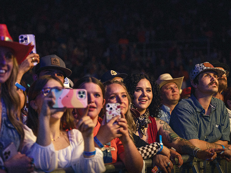 A crowd of people at a concert or event, standing close together and holding up their smartphones to take photos or videos. The audience members are wearing casual clothing, including hats and wristbands, and appear to be enjoying the performance. 
