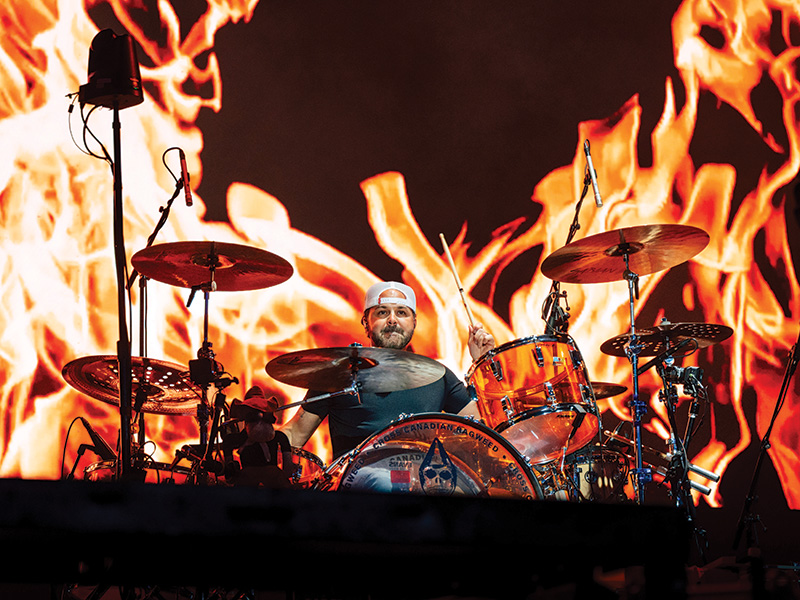A drummer is performing on stage with a backdrop of large, animated flames. The drummer is surrounded by a drum set that includes cymbals and various drums. The intense flames in the background create a dramatic and energetic atmosphere.