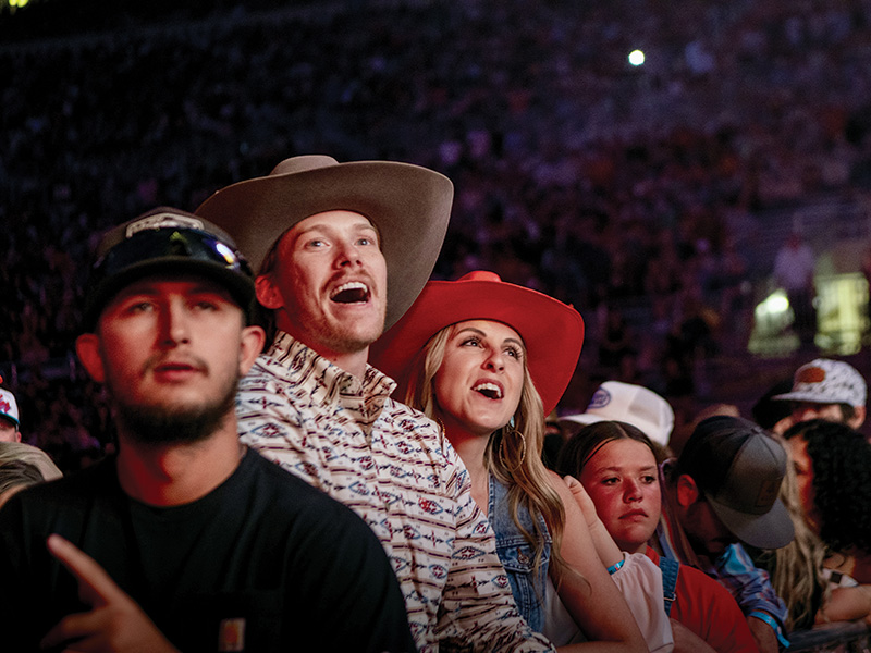 A group of people are standing in a crowded venue. The individuals in the foreground are wearing cowboy hats and casual clothing. The background is filled with many other attendees, creating a lively atmosphere.