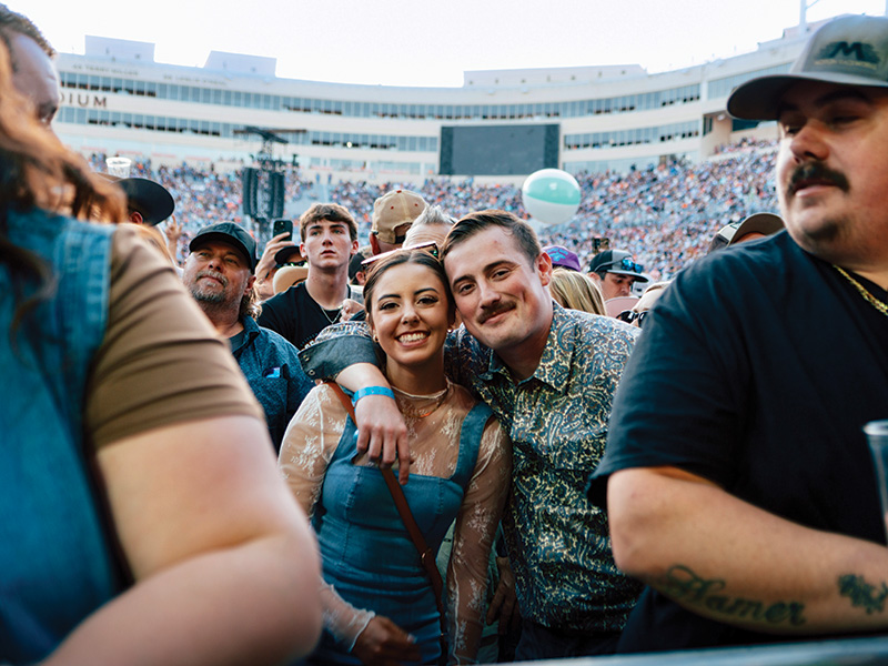 A large crowd of people at an outdoor concert or event, with a stadium full of spectators in the background. The focus is on a group of individuals near the front. One person is wearing a blue dress and has an arm around another person who is wearing a patterned shirt. 