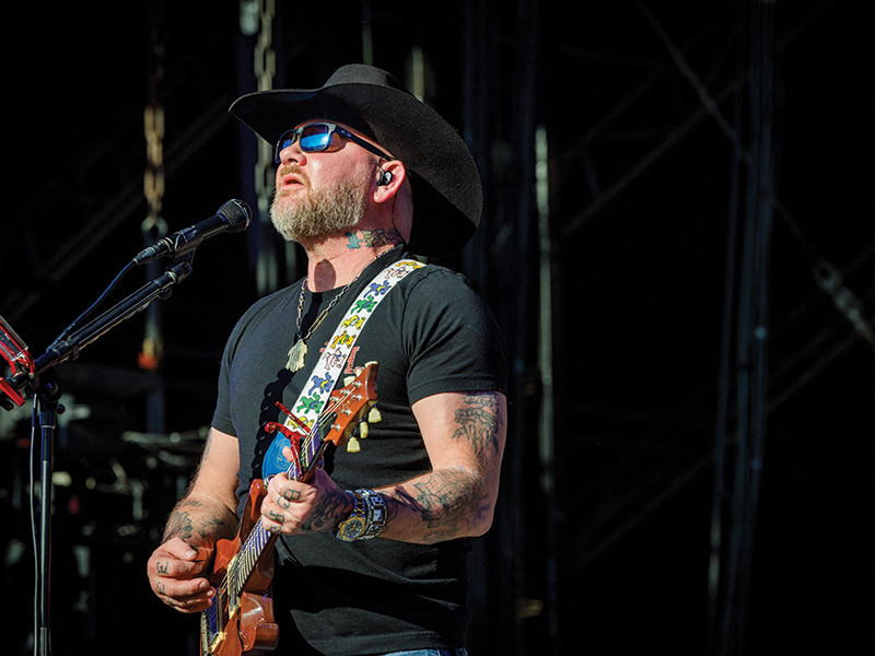 A man with a black cowboy hat, black t-shirt, and tattoos on both arms is performing on stage. The individual is playing an acoustic guitar with a colorful strap and singing into a microphone. 