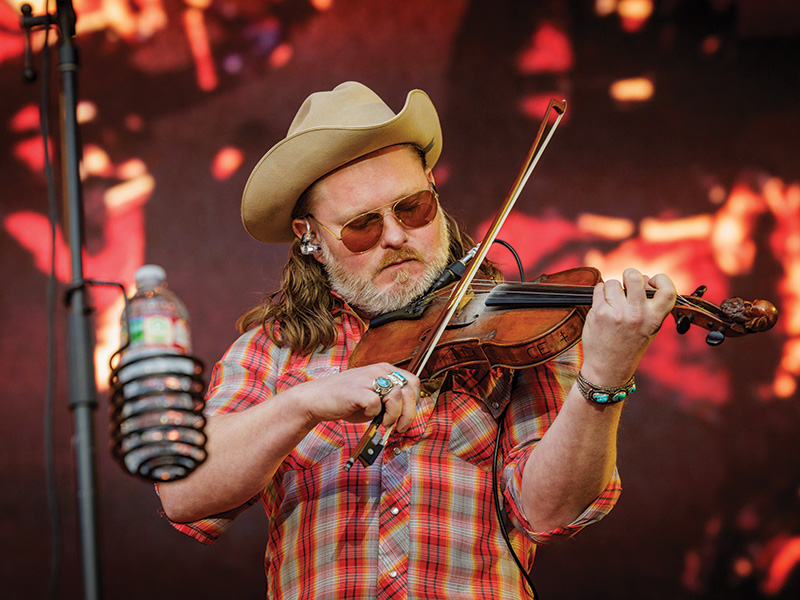 A man wearing a cowboy hat and a plaid shirt is playing a violin. The person has long hair and is wearing rings on the fingers, as well as a bracelet on the wrist. There is a microphone in front of the person, and the background appears to be an out-of-focus stage with warm lighting.