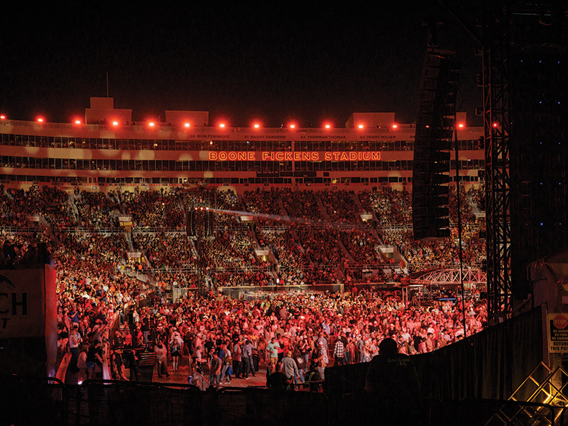 A large crowd of people gathered at Boone Pickens Stadium during nighttime. The stadium is illuminated with red lights, and the stands are filled with spectators.