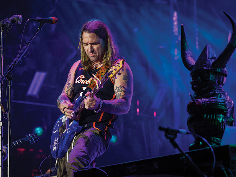 A man with long hair, wearing a sleeveless shirt and tattoos on both arms, is playing an electric guitar on stage. The stage is lit with blue and purple lights, and there is a microphone stand to the left. To the right of the man, there is a statue or prop resembling a horned figure.