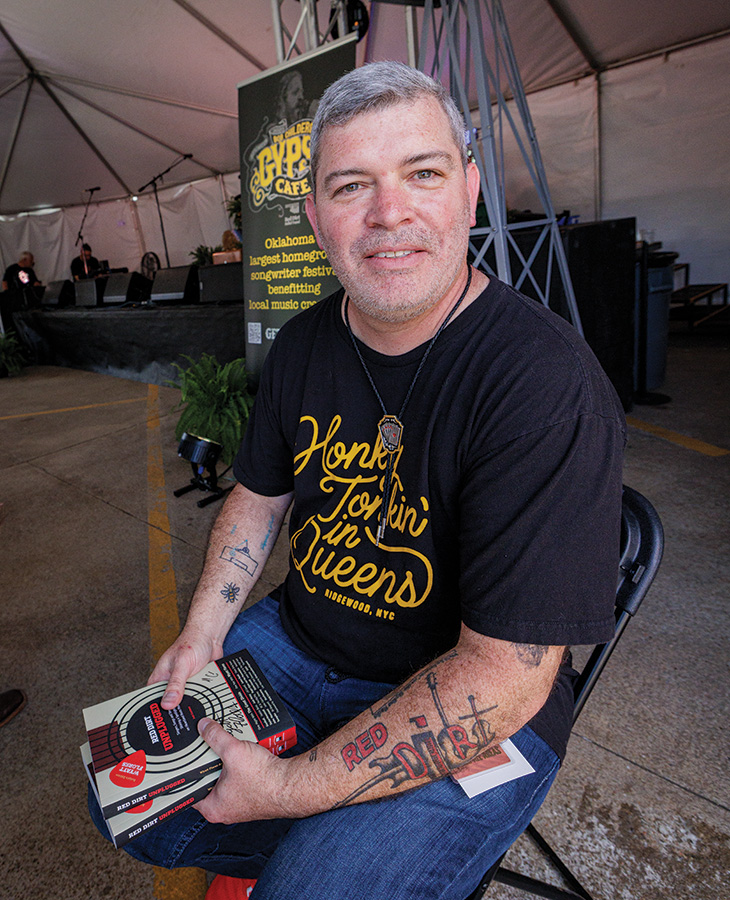 A man with short gray hair is sitting on a black chair under a tent. The person is wearing a black t-shirt with yellow text that reads "Honky Tonkin' in Queens" and holding two books. The person's arms have several tattoos.