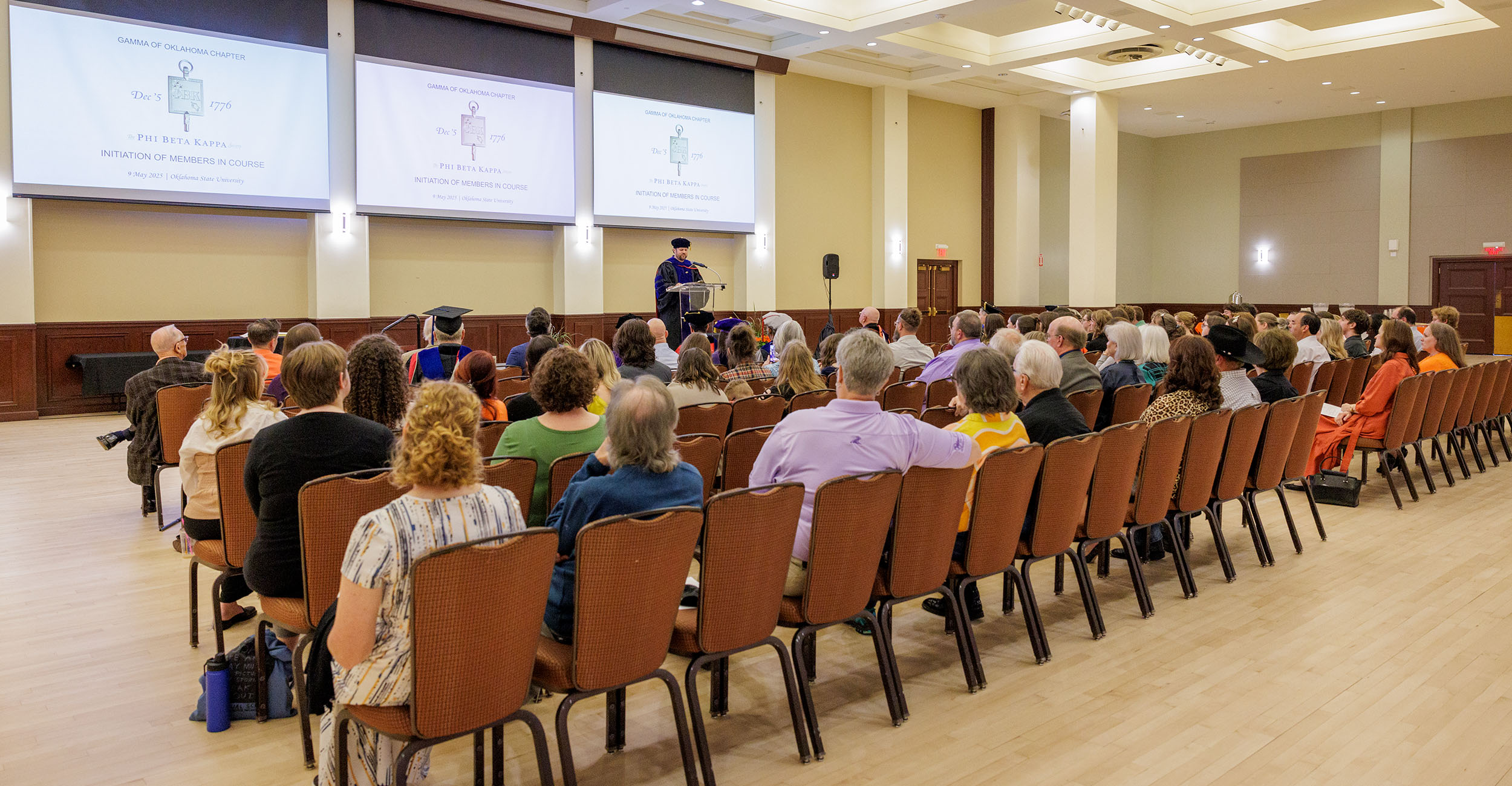 Audience seated in rows inside a large, well-lit hall watching a person in academic regalia speaking at a podium, with presentation slides projected on three screens at the front.