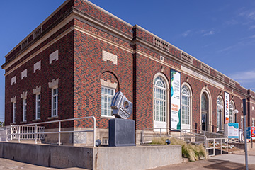 Exterior view of the red‑brick Postal Plaza Gallery building, showing arched windows, exhibition banners, an accessible ramp, and a metal sculpture at the entrance on a sunny day.