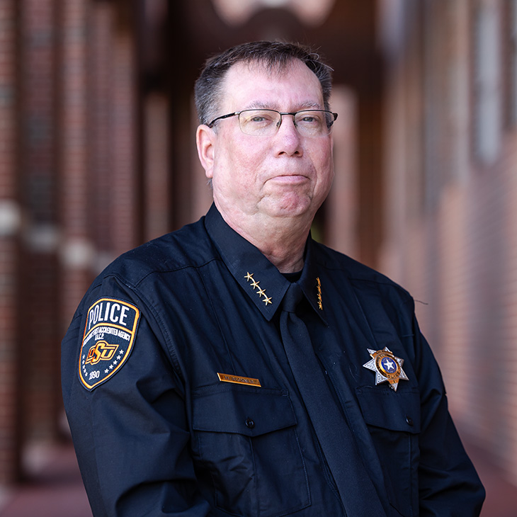 Person in a black police uniform with an OSU Police shoulder patch stands outdoors in front of a brick walkway, posed for a formal portrait.