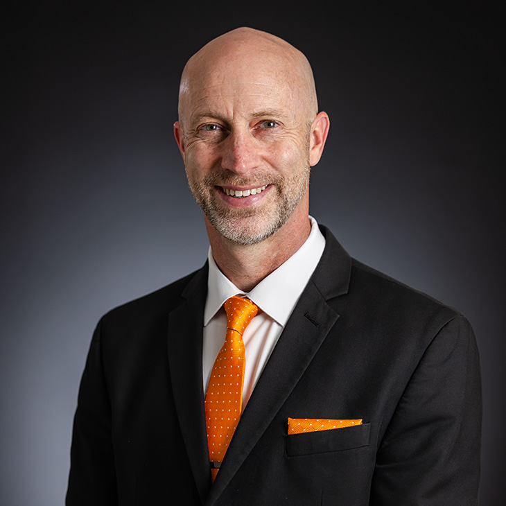 Person wearing a black suit jacket with a white dress shirt, an orange polka-dot tie, and a matching orange pocket square, posed against a dark studio background.