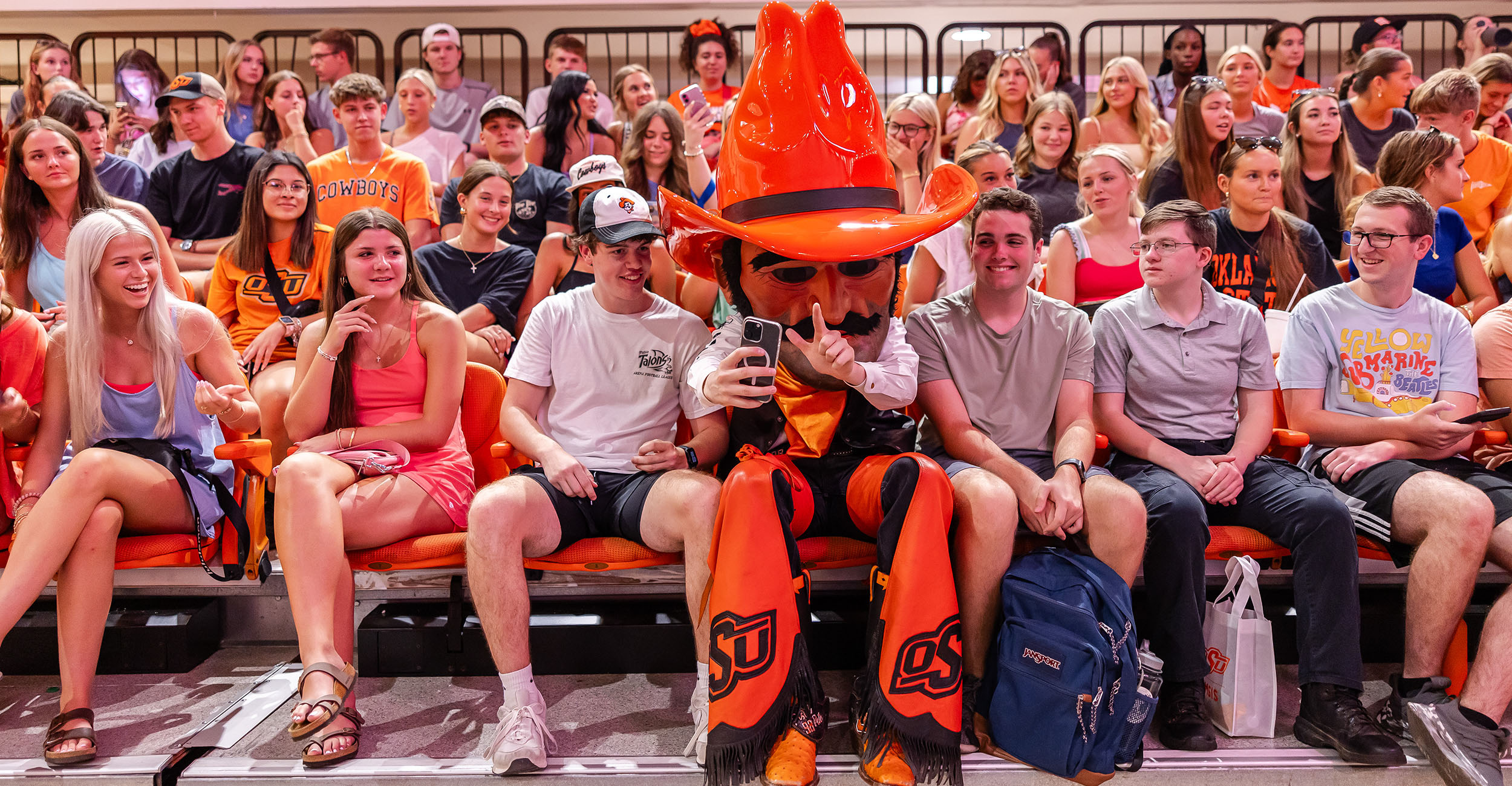A group of people sitting in stadium seats, many wearing orange and black. At the center, a person in an orange mascot costume with a large cowboy hat and chaps labeled 'OSU'. The crowd in the background is also seated and facing forward.