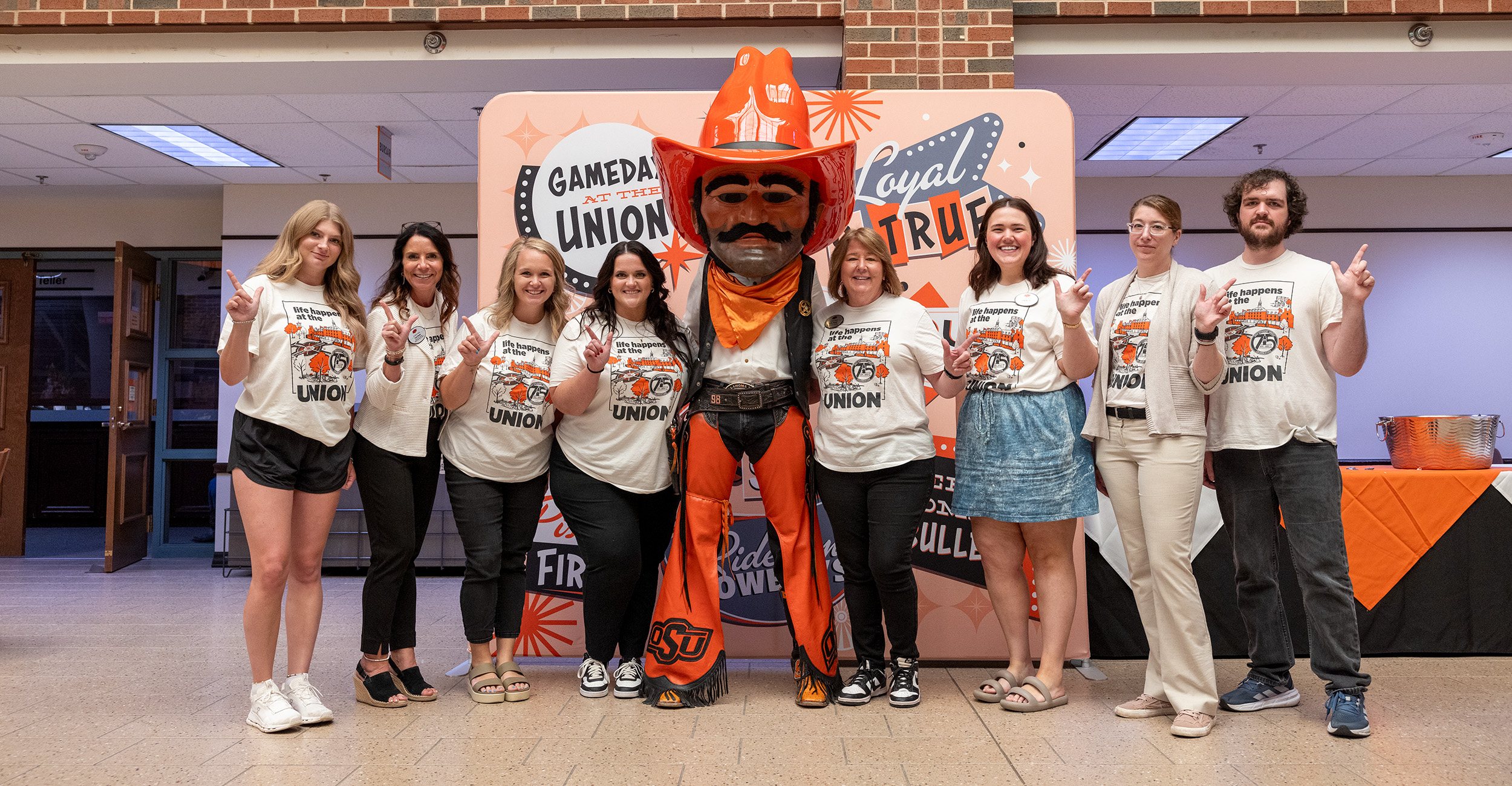 A group of attendees pose with a large costumed mascot in a campus building lobby during a Student Union anniversary kickoff event, with event tables and signage visible in the background.