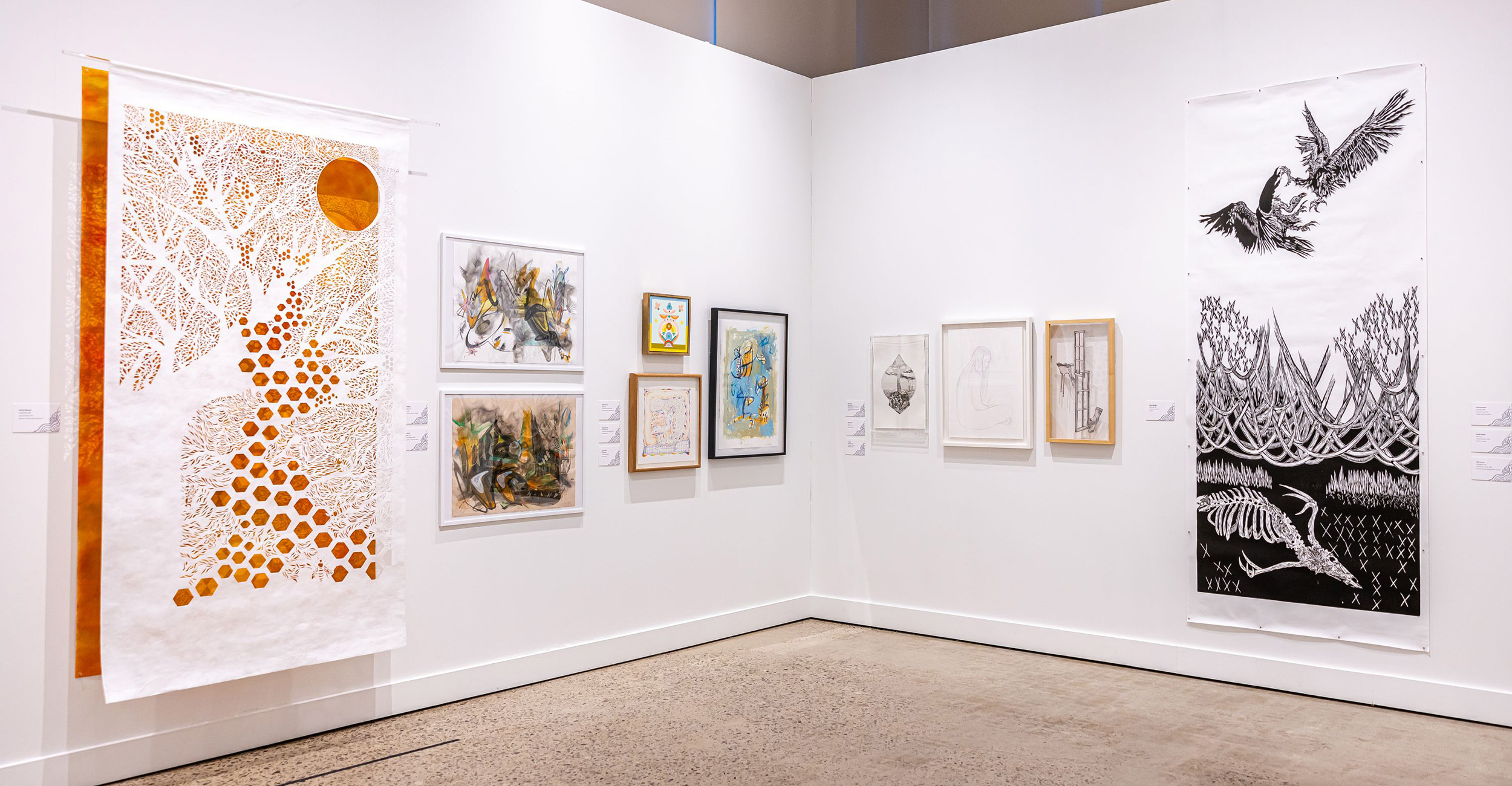 An art gallery corner with white walls displaying various artworks. On the left, a large piece features an orange sun and tree-like design with circular patterns. Several framed drawings and paintings are arranged in the center, and on the right, a tall black-and-white piece shows birds flying above abstract shapes.