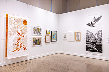 An art gallery corner with white walls displaying various artworks. On the left, a large piece features an orange sun and tree-like design with circular patterns. Several framed drawings and paintings are arranged in the center, and on the right, a