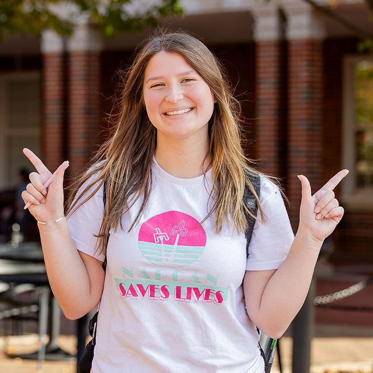 A smiling college student stands outside on campus, holding up both hands in OSU’s “pistols firing” gesture. She wears a white T-shirt that says “Narcan Saves Lives” with a pink and turquoise design and has a backpack on her shoulders.