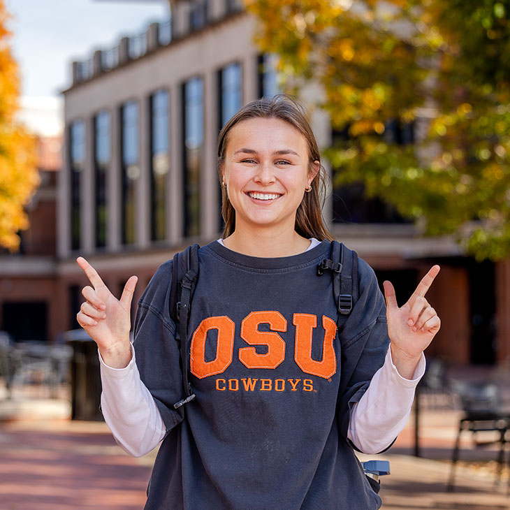 Student smiling and posing with OSU hand signs while wearing an “OSU Cowboys” shirt on campus, with fall trees and an academic building in the background.