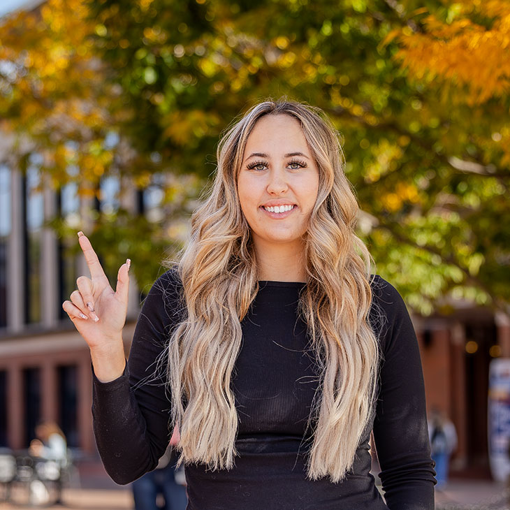 Student smiling and holding up an OSU hand sign while standing on campus in front of fall trees and an academic building.