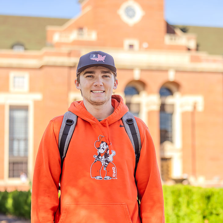 A smiling college student stands in front of a red brick campus building wearing an orange OSU hoodie, gray backpack, and dark cap.