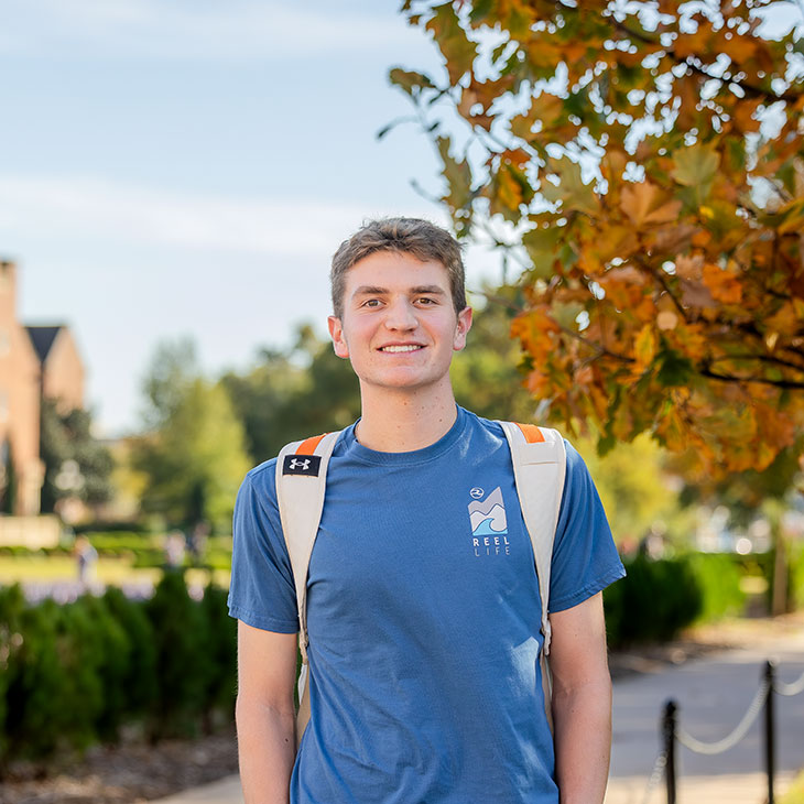 Student wearing a blue “Reel Life” T-shirt and backpack smiles on campus with fall foliage in the background.