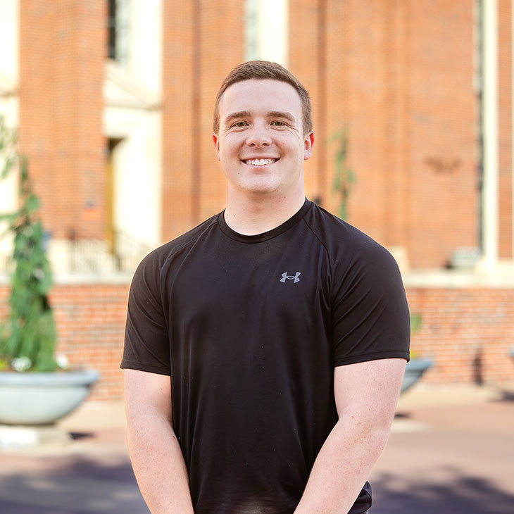 A smiling college student stands outside a red brick building wearing a black Under Armour T-shirt.