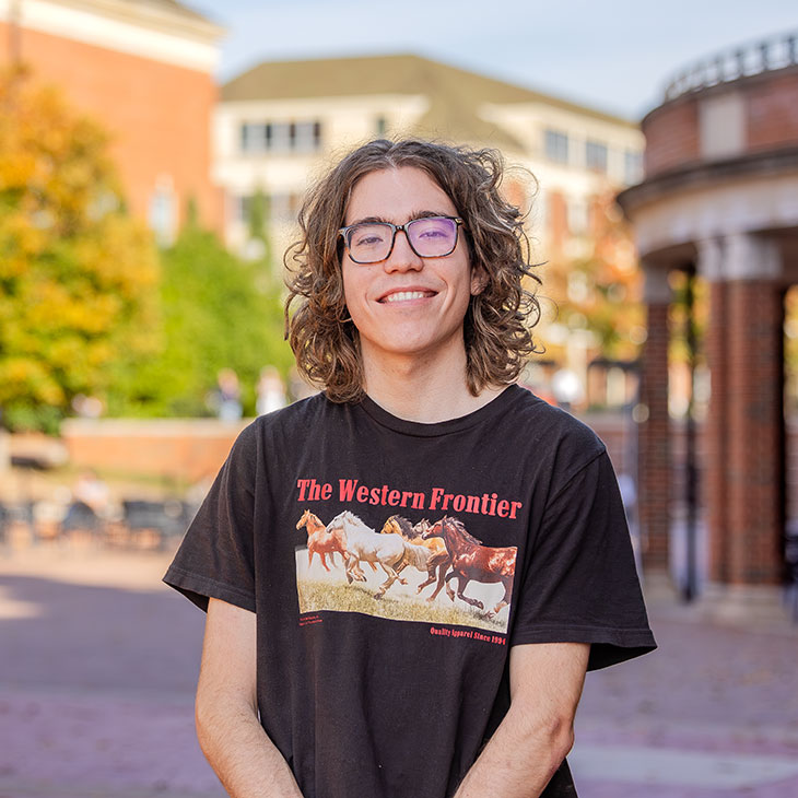 Student wearing a black “The Western Frontier” T-shirt smiles on campus with fall colors and brick buildings in the background.