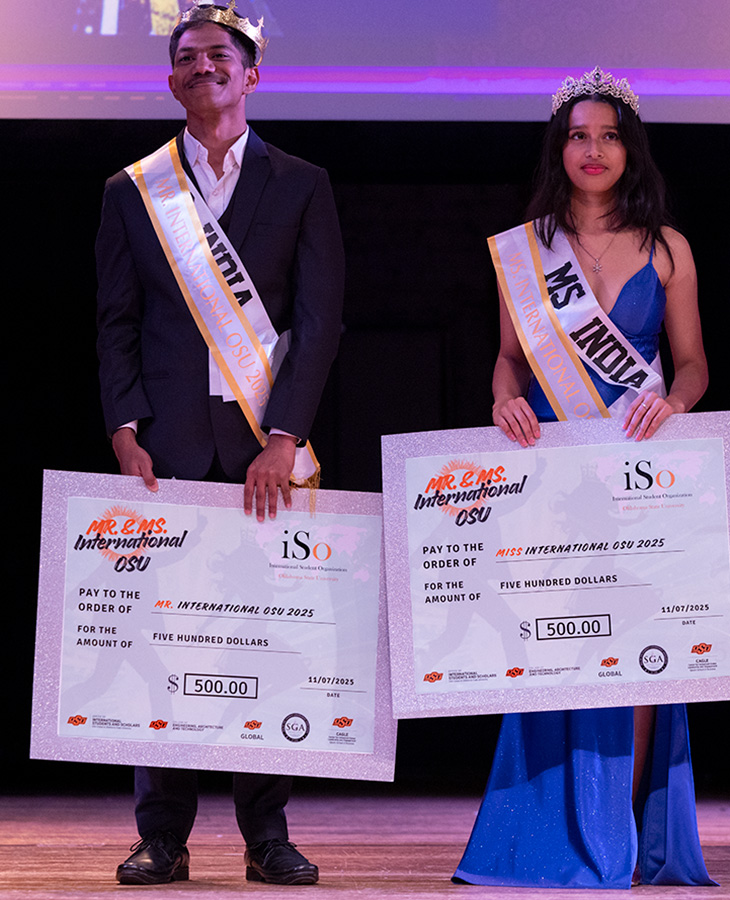 Two individuals on stage wearing sashes labeled ‘Mr. India’ and ‘Ms. India,’ each holding a large ceremonial check for $500 with the text ‘Mr. & Ms. International OSU’ and event details.