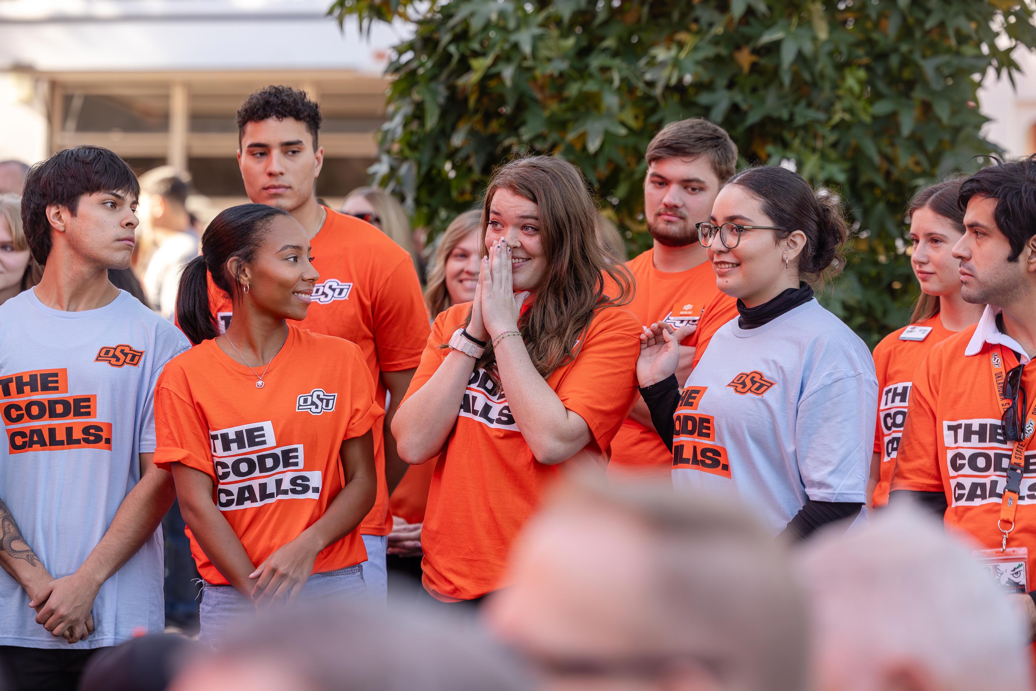 A young woman in an orange OSU “The Code Calls” T-shirt stands at the center of a group of students, holding her hands to her face in an emotional moment. Other students around her smile and look on during the outdoor event.