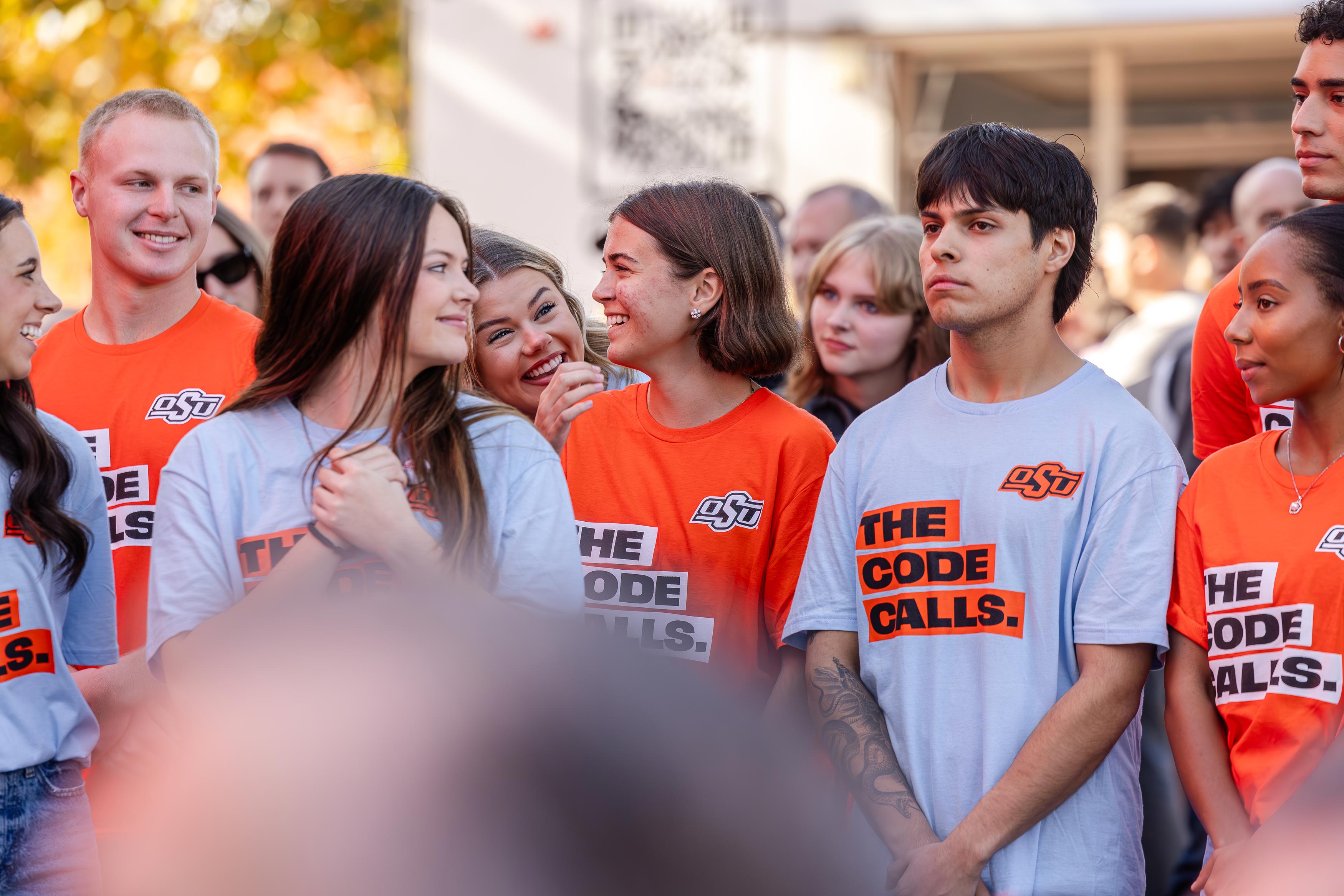 A group of OSU students wearing orange and light-blue “The Code Calls” T-shirts stand together outside, smiling and talking with one another. Autumn trees and a crowd are visible in the background.