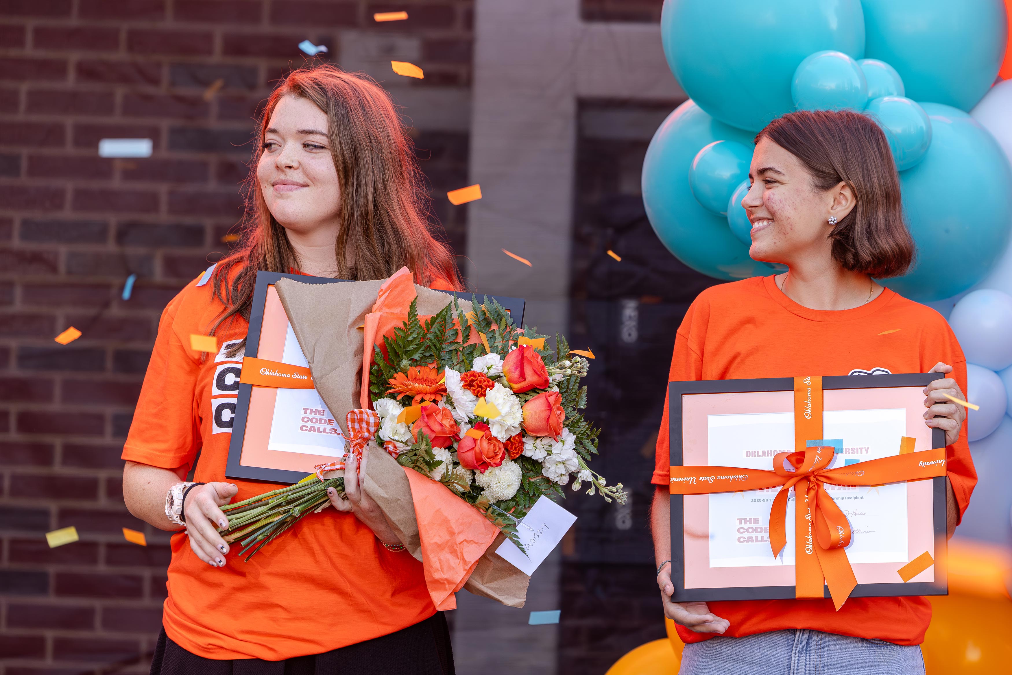 Two young women wearing bright orange OSU shirts smile while holding framed certificates and bouquets during an outdoor celebrati