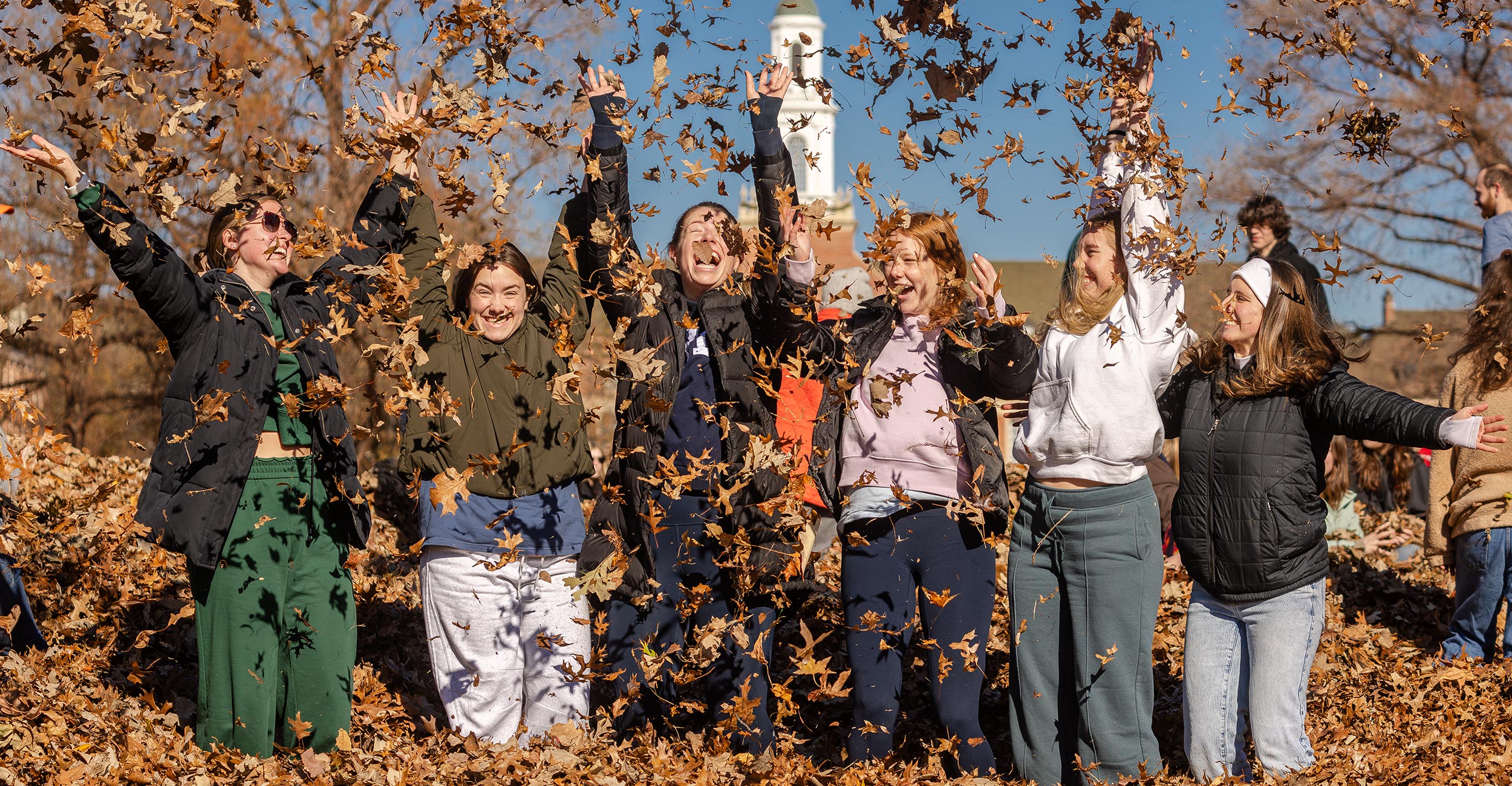 a group of college students stand in a leaf pile and throw leaves in the air
