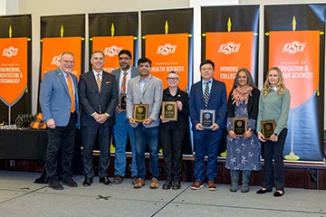 A group photo of 6 recipients holding award plaques on a stage.