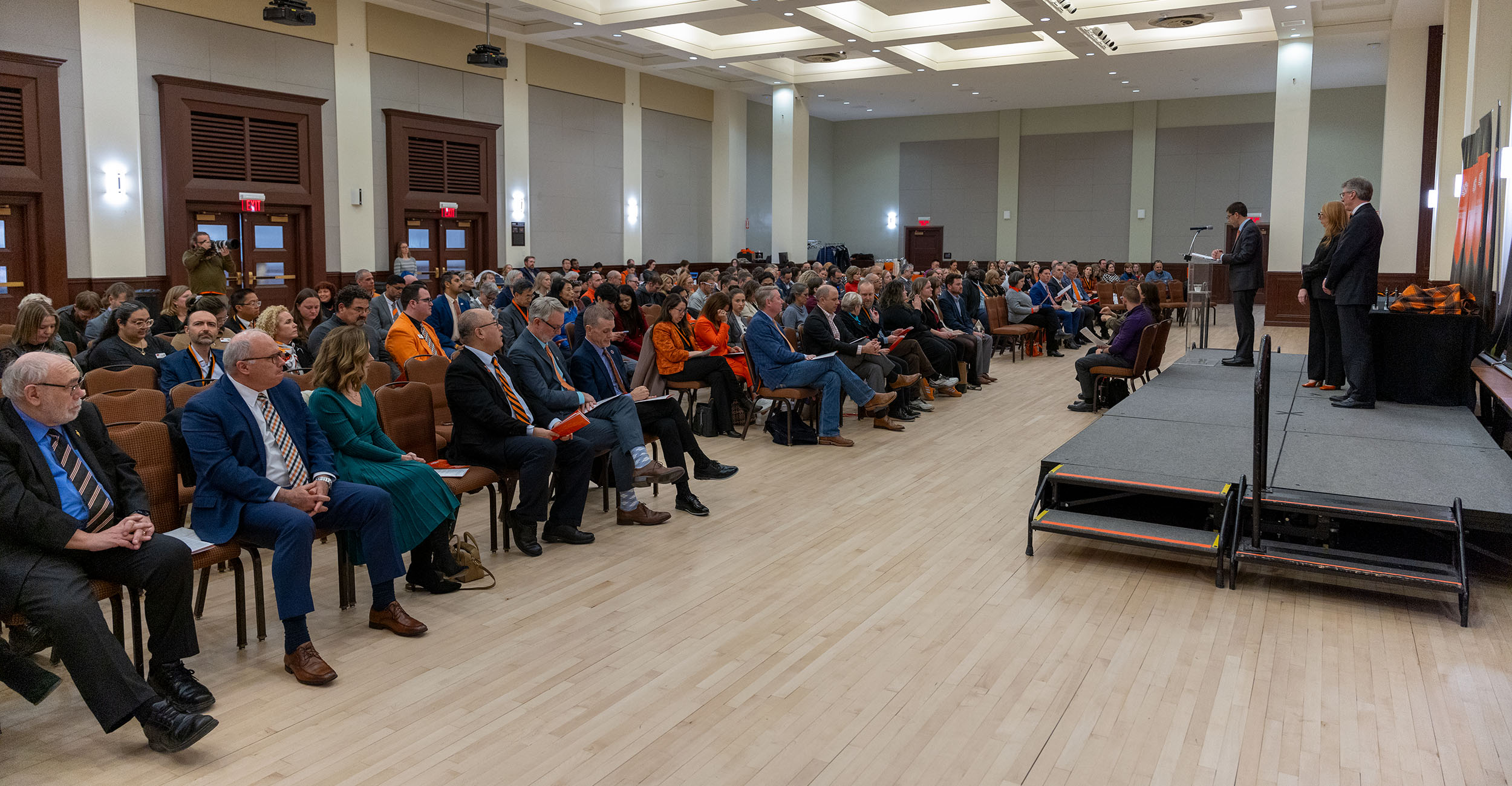 Large audience seated in a spacious hall facing a stage where several people stand near a podium. Oklahoma State University banners are displayed behind the stage.