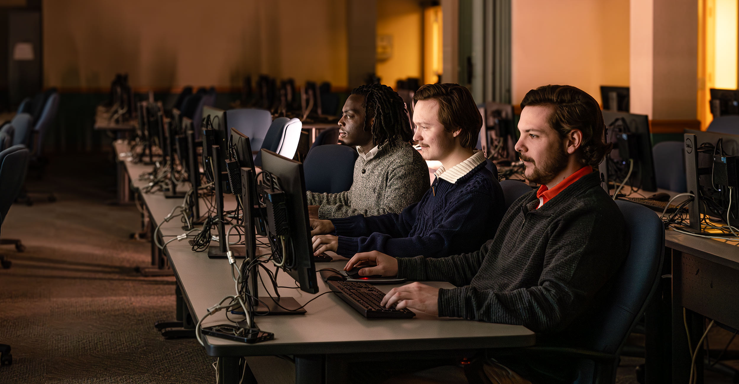 Three male students sit next to each other in a computer lab, looking at their screens, working on data files.