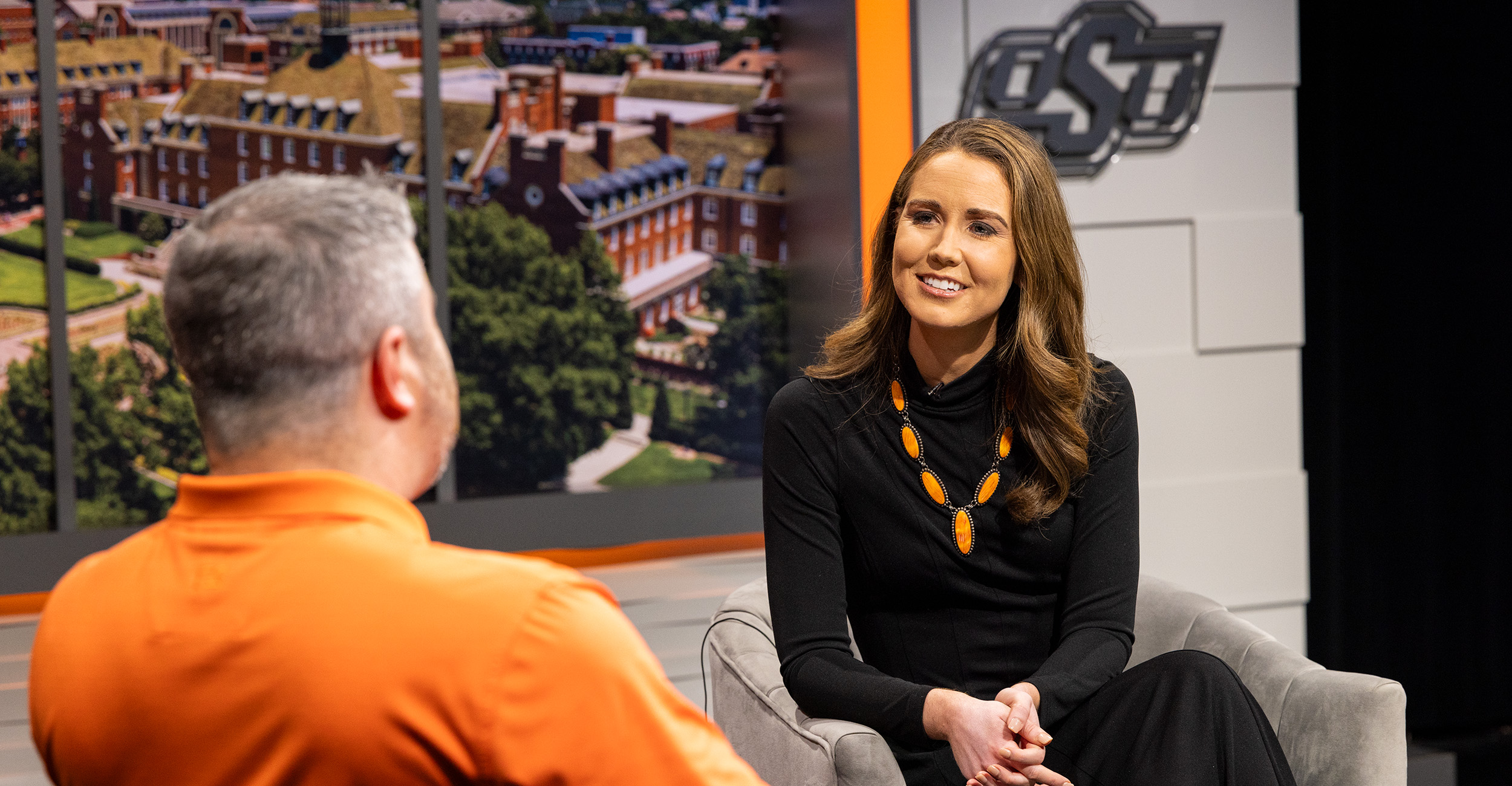 Two people sit across from each other in a studio setting, with one wearing an orange shirt and gesturing while speaking, and the other dressed in black with a gold necklace, seated against a backdrop featuring OSU campus imagery and the OSU logo.
