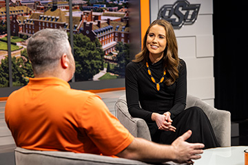 Two people sit across from each other in a studio setting, with one wearing an orange shirt and gesturing while speaking, and the other dressed in black with a gold necklace, seated against a backdrop featuring OSU campus imagery and the OSU logo.